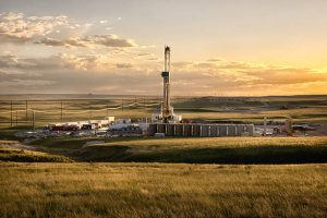 A lone drilling Fracking Rig set against the dramatic landscape of the American Prairie lands. The sun is setting just off camera creating beautiful yellows and oranges on the clouds above. In the far distance, wind turbines are visible.